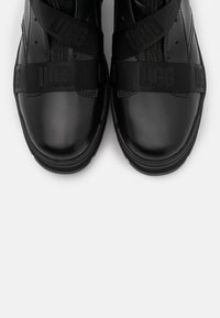 Mustat nahkakengät, joissa on sileä pinta, joissa on ristikkäin mustat hihnat, joissa on logo, pyöreä kärki ja kumipohja.