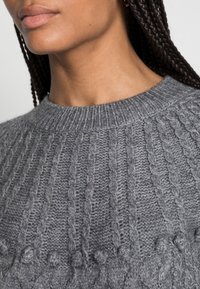 Detailní záběr osoby v šedém svetr s copánkovými vlasy viditelnými na pravé straně.