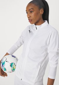 White athletic jacket with a zippered front, black logo, and mesh side panels. A colorful patterned soccer ball is held in one hand.