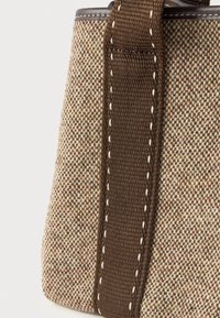 Κοντινή λήψη από μπεζ και καφέ υφασμάτινη τσάντα με σκούρα καφέ λουρί, που έχει λευκή ραφή κατά μήκος των ακμών.