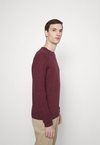 Burgunderfarbener Wollpullover mit Rundhalsausschnitt, gerippten Bündchen und Saum, getragen mit beigefarbenen Hosen, vor einem neutralen Hintergrund platziert.