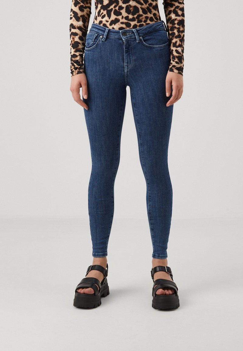 Jeans ajustados de denim en azul oscuro, con un diseño clásico de cinco bolsillos y una textura suave, combinados con sandalias negras gruesas.