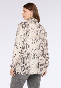 Donna con capelli castani che indossa grandi orecchini a cerchio e una camicia a maniche lunghe con stampa a serpente leggera, abbinata a jeans grigi.