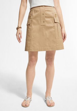 Person iført en beige A-formet nederdel med knapper foran og klaplommer, en hvid top, armbånd og hvide sandaler med åben tå.