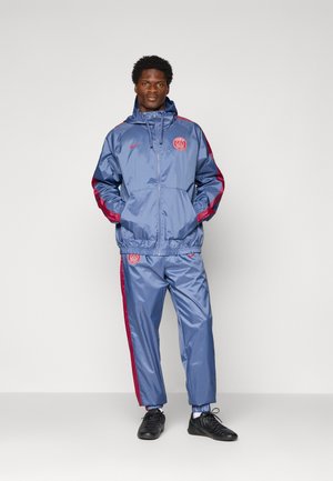 Man wearing blue and red Paris Saint-Germain tracksuit with jacket hood up, standing with hands in pockets against plain background.