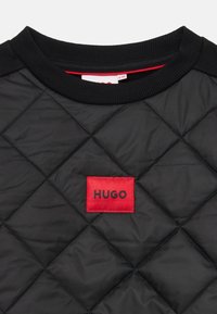 Felpa nera trapuntata con colletto a costine, caratterizzata da una patch rettangolare rossa con il logo "HUGO" in nero. Superficie testurizzata con motivo a diamante.