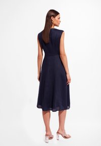 Granatowa lniana sukienka bez rękawów z dopasowaną górą i rozkloszowanym dołem. Tył ma gładką teksturę i subtelne szwy. Przezroczyste sandały na obcasie.