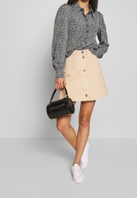 Black floral print blouse with puffed sleeves, paired with a beige denim skirt featuring front buttons. Holding a black handbag, with white sneakers.