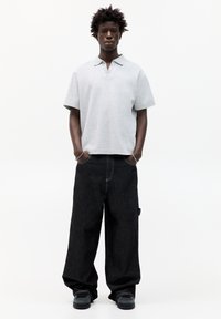 Grå kortærmet polo med krave, parret med løse, mørke denim jeans. Modellen står med hænderne i lommerne og bærer sorte sneakers.