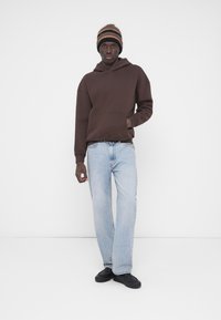 Καφέ φούτερ με κουκούλα και τσέπη μπροστά, ανοιχτό μπλε τζιν με ίσιο κόψιμο και μαύρα παπούτσια, συνδυασμένα με ριγέ πλεκτό σκουφί.