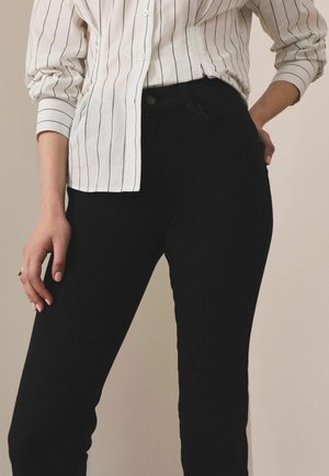 Inimene, kes kannab valget pika varrukaga särki mustade vertikaalsete triipudega ja kõrge vöökohaga musti pükse, käsi toetub puusale ja reiele.