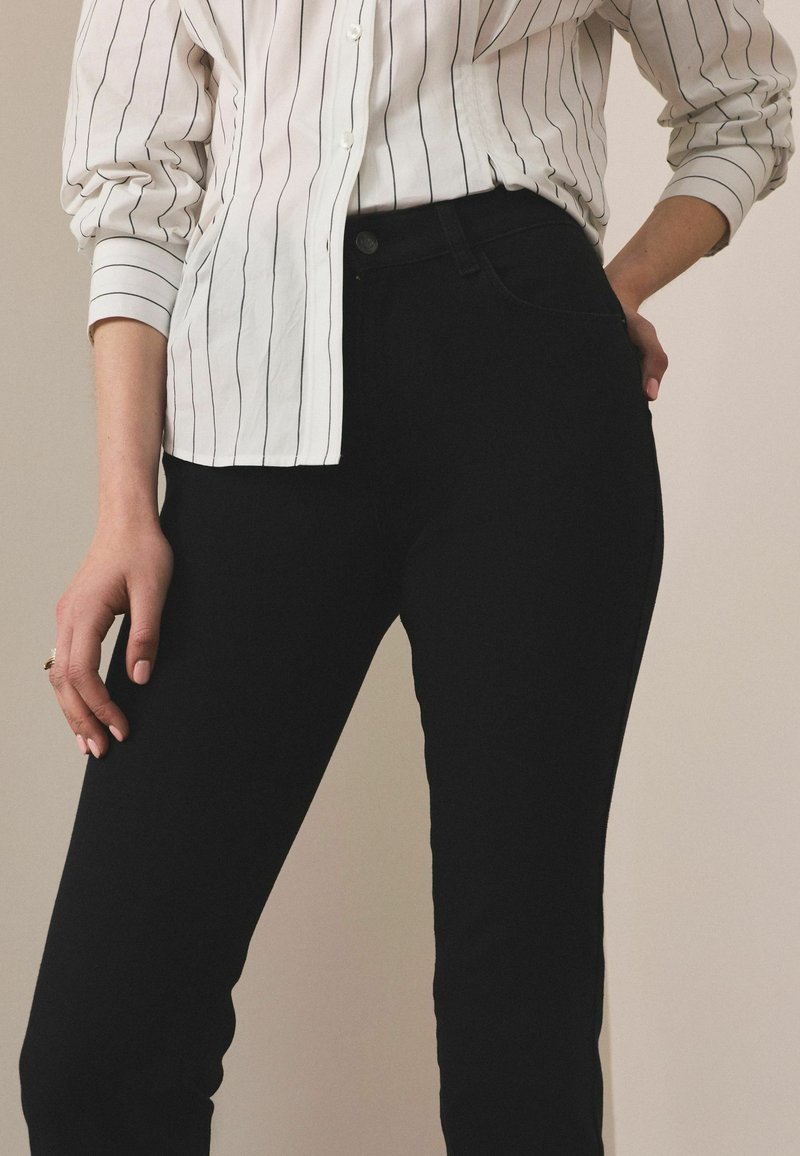 Persona con camisa blanca de manga larga con rayas verticales negras y pantalones negros de tiro alto, con la mano apoyada en la cadera y el muslo.