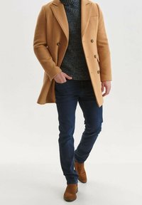 Manteau en mélange de laine marron clair à double boutonnage, pull marine, jean foncé et bottines en daim marron. Fond minimaliste.