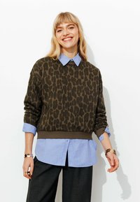 Pull imprimé léopard vert olive superposé à une chemise bleu clair à boutons, avec des manches retroussées et un ourlet arrondi.