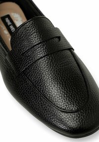 Mocassino in pelle nera con finitura testurizzata, dotato di tomaia rialzata e punta liscia. Forma arrotondata, design slip-on e dettagli di cucitura minimi.
