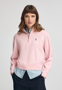 Ljusrosa zip-up tröja med långa ärmar, ribbad fåll och en liten svart logotyp. Bärs över en blå denimskjorta. Mjuk textur.
