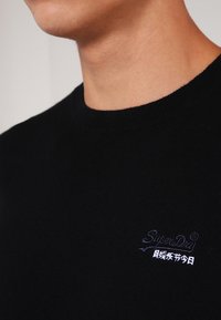 Czarny sweter dzianinowy z okrągłym dekoltem, z małym haftowanym logo w kolorze fioletowym na lewej stronie klatki piersiowej. Gładka struktura, casualowy design.
