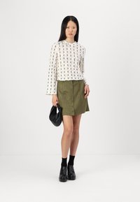 Fehér mintás, hosszú ujjú blúz olíva zöld velúr szoknyával, amely elöl gombos; a modell fekete táskát tart, fekete cipőt és zoknit visel.