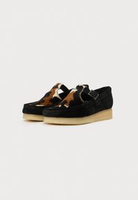 Fekete velúr cipők narancssárga és barna tehénmintás kiegészítőkkel, slip-on dizájnnal és világos színű gumi talppal.