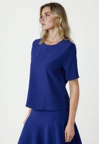 Kobieta o blond włosach, ubrana w prosty niebieski top z krótkim rękawem i dopasowaną spódnicę, patrząca w bok na jednolitym białym tle.