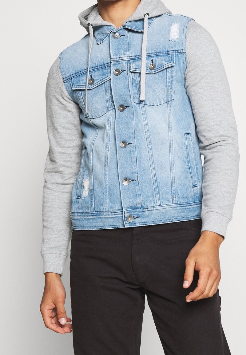 Chaqueta de mezclilla con frente azul claro, mangas de sudadera gris, dos bolsillos en el pecho, botones de metal y detalles desgastados. Combinada con pantalones negros.