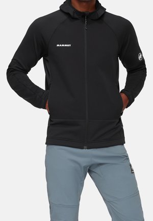 Mand iført sort hættejakke med lynlås og "Mammut"-logo på brystet og venstre arm, kombineret med lyseblå bukser. Hænderne er i jakkens lommer.