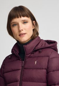 Burgundowa kurtka puchowa z zamkiem błyskawicznym, z kapturem, pikowaną teksturą i subtelnym logo na piersi. Nosi ją na czarnym golfie.