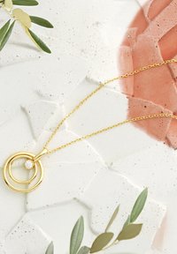 Guldhalsband med två sammanlänkade cirkulära hängen, med en liten kristallaccent, på en delikat kedja mot en texturerad bakgrund.