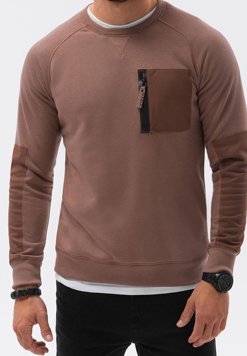 Homme portant un sweat-shirt marron à manches longues avec une poche poitrine zippée et des coudières, un pantalon noir, une montre et un bracelet.
