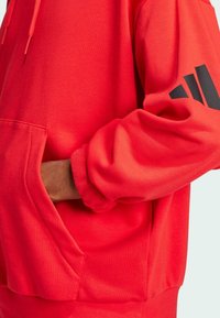 Röd hoodie i bomullsblandning, med en framficka och svart logodetalj på ärmen. Mjuk textur och avslappnad passform.