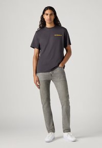 Sötétszürke pamut póló sárga "Levi Strauss & Co." logóval, világosszürke farmernadrággal és fehér sportcipővel párosítva. A modell frontális pózban áll.