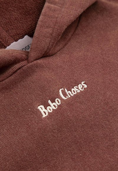 Nahaufnahme eines gefalteten braunen Stoffes mit weißem, gesticktem Schriftzug "Bobo Choses" und teilweise sichtbarem Kleidungsetikett im Kragen.