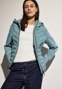 Giacca turchese chiaro con frontale imbottito, tasche con zip e cappuccio, abbinata a una maglietta bianca e pantaloni in denim blu scuro.