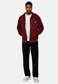 Sudadera con cremallera color marrón burdeos y cordones, camiseta blanca debajo, pantalones de chándal negros, zapatillas gris y detalle sutil del logo en la sudadera.