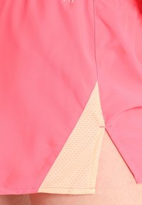 Los pantalones cortos rosas cuentan con una abertura lateral que revela un forro de malla beige claro con un patrón texturizado. Están fabricados con una tela ligera para mayor transpirabilidad.