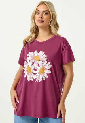Lyshåret kvinde iført en løs magenta T-shirt med tre store hvide margueritter med gule centre trykt på forsiden.