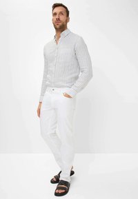 Langärmliges, knöpfbares Hemd in Hellgrau mit weißen vertikalen Streifen, kombiniert mit weißen Hosen und schwarzen Slipper-Sandalen. Schlichtes, modernes Design.