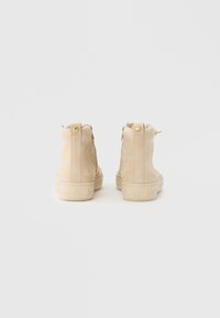 Μπεζ μποτάκια αστραγάλου από σουέντ με υφή, με χρυσό κουμπί και φερμουάρ πίσω, τοποθετημένα σε ανοιχτόχρωμη λαστιχένια σόλα.