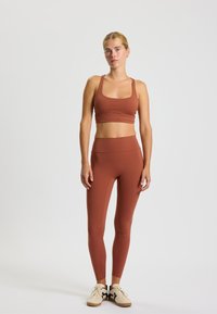 Bruine sportbeha en high-waisted legging gemaakt van rekbaar materiaal. Draagt samen met beigerode sneakers, met een vetersluiting en donkere accenten.
