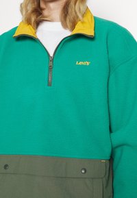 Pletený pulovr v teal barvě se zipovým límcem, kontrastním žlutým vnitřním límcem, zeleným spodním panelem a vyšitým oranžovým logem.