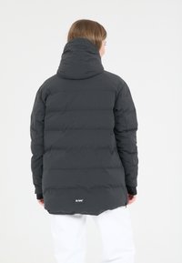 Mörkgrå pufferjacka med huva, med kviltade sektioner och en logo vid nederkanten. Materialet verkar mjukt och vadderat.