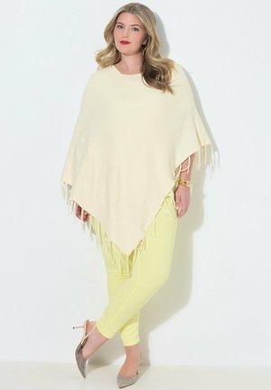 Vrouw met golvend blond haar, gekleed in een crèmekleurige poncho met franjes, gele broek, gouden armband en oorbellen, en beige hakken, staand tegen een witte muur.