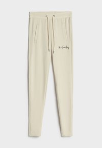 Krémové jogger kalhoty vyrobené z měkké tkaniny, s elastickým pasem na šňůrku, bočními kapsami a vyšitým textem "Ty někdy."