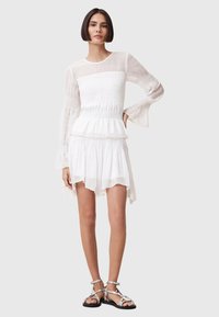 White dress featuring a smocked bodice, flared sleeves, and layered skirt. The fabric is lightweight with a sheer overlay. Paired with silver sandals.