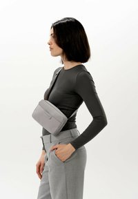  Šedá crossbody taška s hladkou texturou, obdélníkovým tvarem, zipovým uzávěrem a nastavitelným popruhem, nošená na černém topu s dlouhým rukávem.