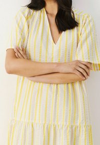 Sukienka w paski z żółtymi i białymi pionowymi liniami, krótki rękaw, dekolt w serek oraz warstwowym dołem. Tkanina wydaje się mieć fakturę i być lekką.