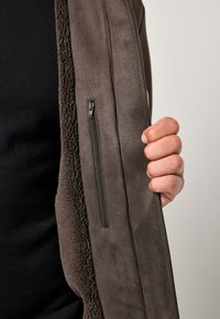 Mouwloos bruin vest met een gladde buitenkant en een nepbont voering. Beschikt over een ritszak aan de linkerkant. Hand die de stof vasthoudt.