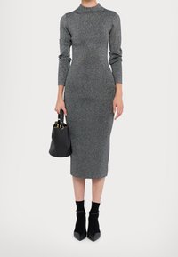 Robe en tricot grise à manches longues, col haut et silhouette ajustée. Assortie à un sac à main noir et des escarpins noirs à talons pointus, avec des chaussettes de cheville.