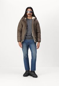 Giacca imbottita marrone con fodera in pile bianco, maglione grigio, jeans blu e sneakers nere con motivo. Gli occhiali da sole riflettenti completano il look.