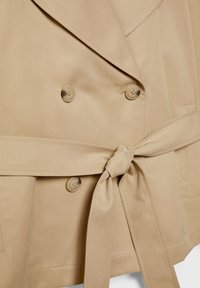Gros plan sur un trench-coat beige montrant de larges revers, quatre boutons et une ceinture en tissu nouée à la taille.